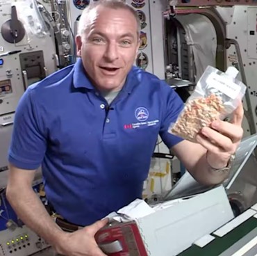 Astronaut David Saint-Jacques aboard the International Space Station, holding a pouch of chili, with technical equipment and compartments in the background.
